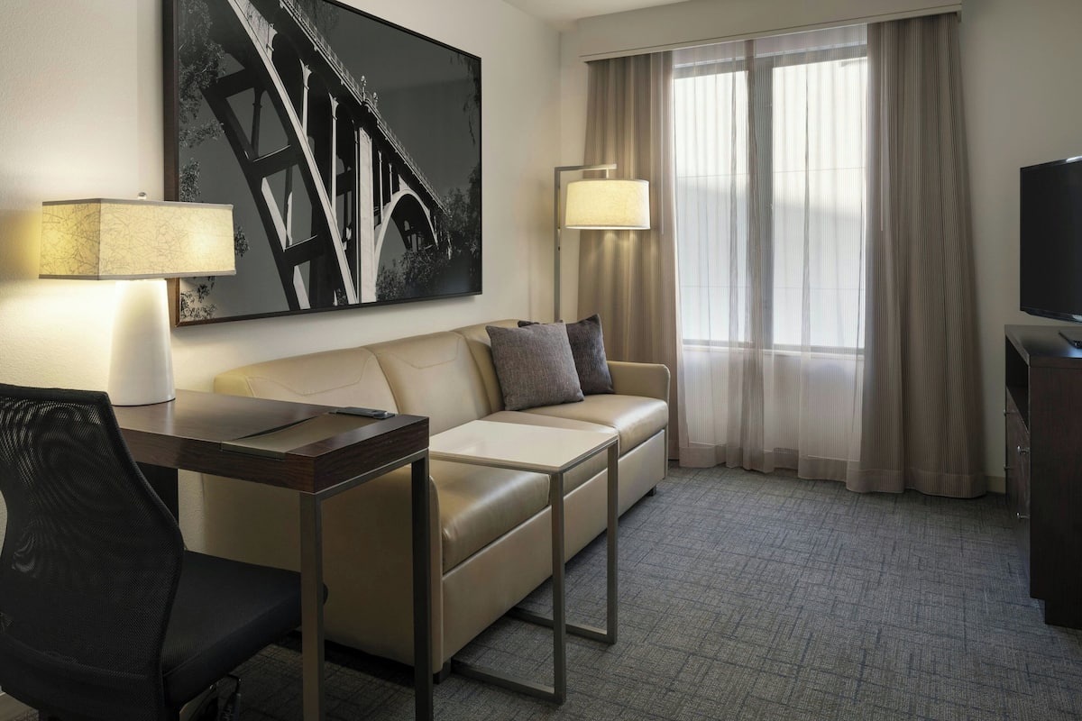 A hotel room with a sofa, a desk with a chair, a TV, a lamp, and a large abstract painting on the wall.