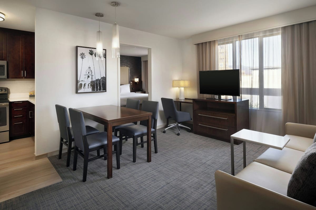 A hotel room with a dining area, a sofa, a TV, a kitchen, and a large window with curtains.