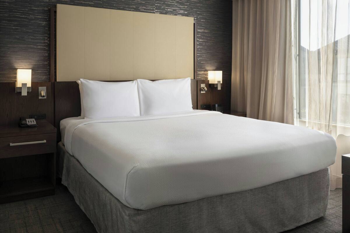 A hotel room with a large bed, a wooden headboard, two bedside lamps, a nightstand with a phone, and a window with sheer curtains.