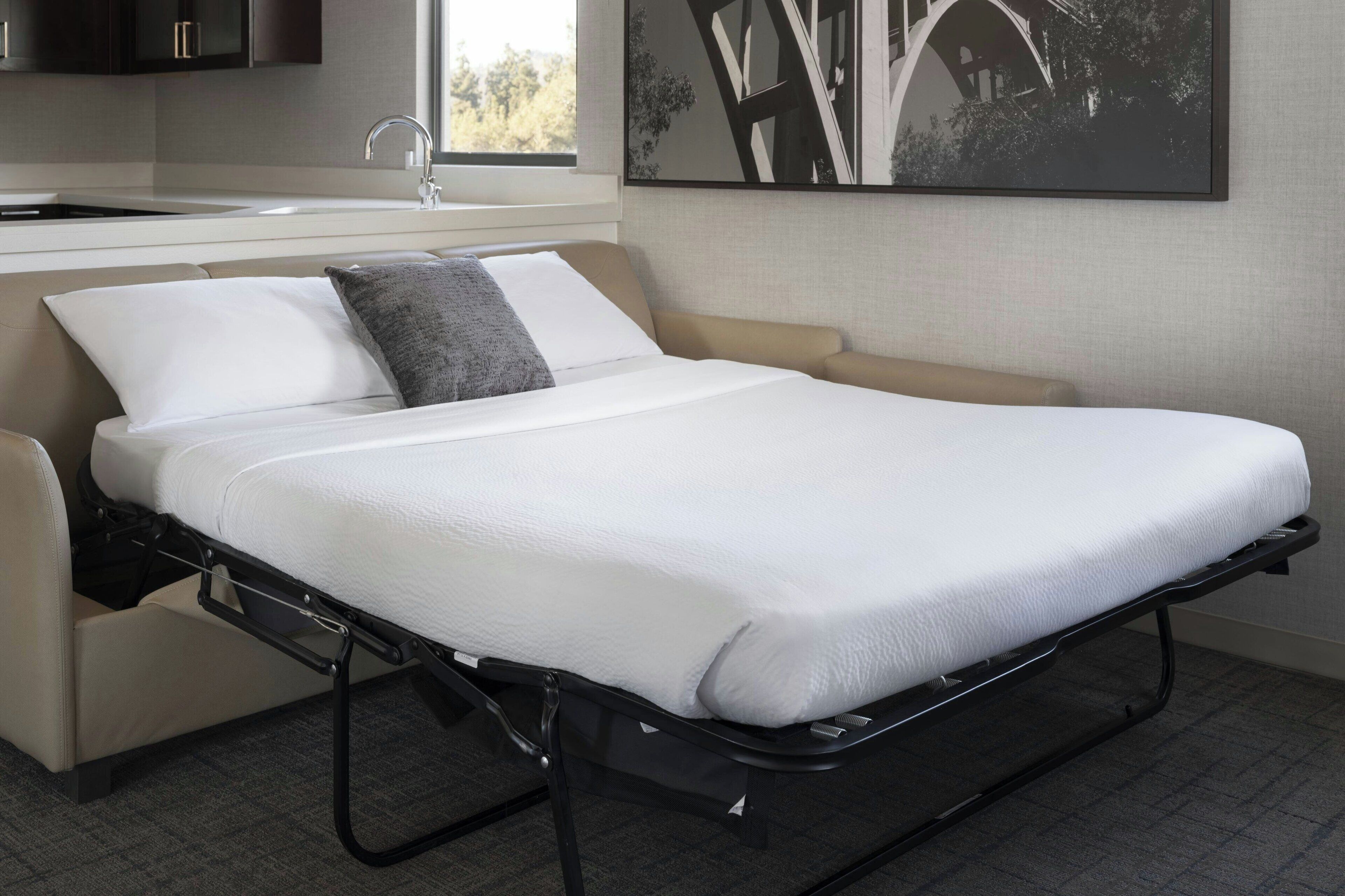 Una cama compacta con colchón blanco y un escritorio plegable, un pequeño sofá, una ventana con vistas a árboles, y una zona de cocina con armarios y un lavamanos.