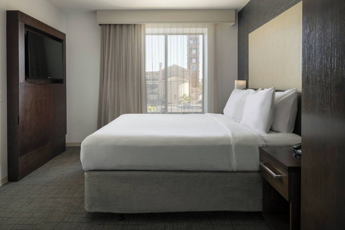A hotel room with a large bed, a flat-screen TV mounted on the wall, a window with curtains, and a view of buildings outside.