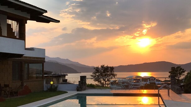 Una casa moderna con piscina, vista al atardecer sobre un lago y montañas al fondo.