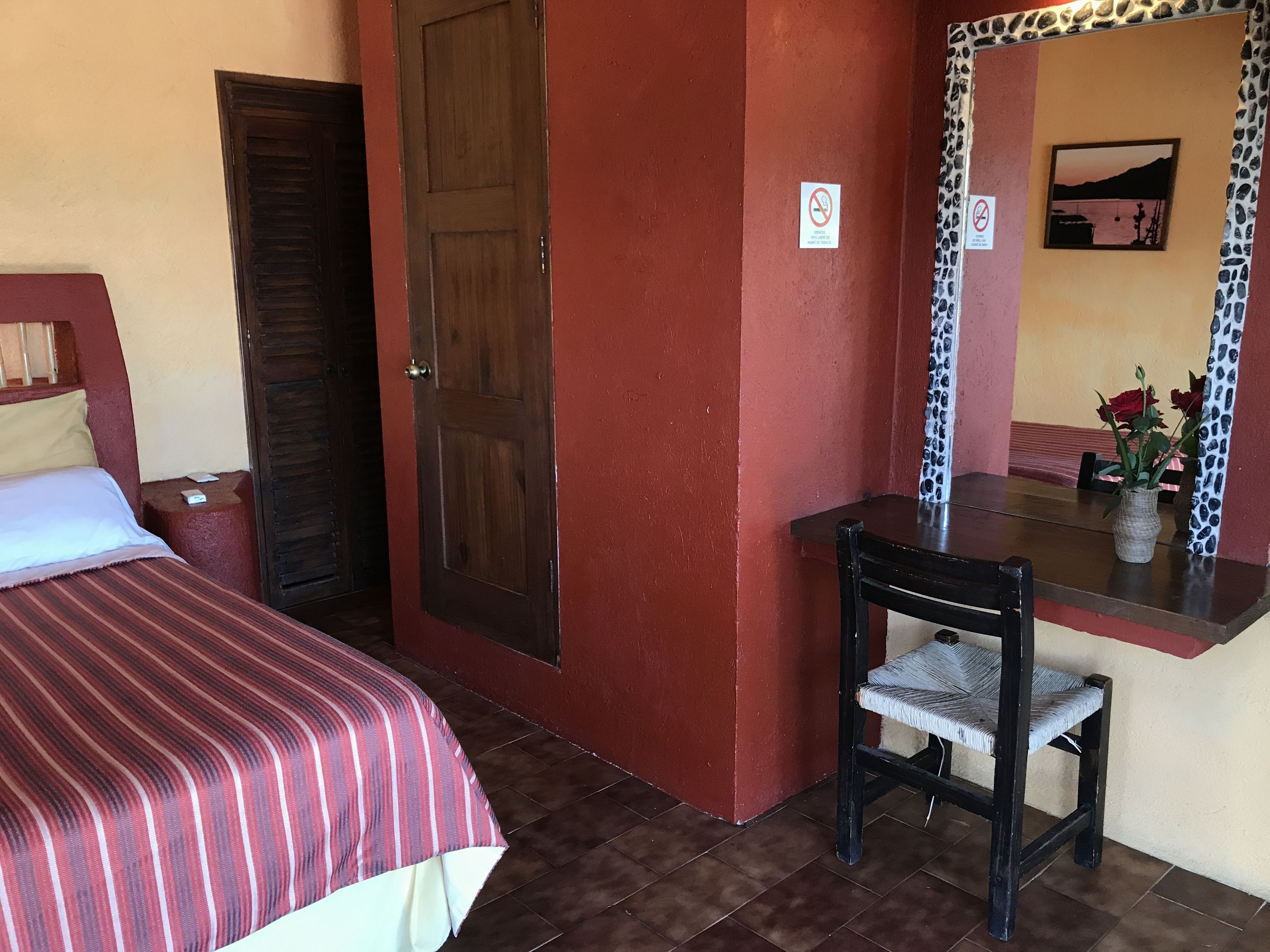 A hotel room with a bed, a desk, and a chair. There is a door and a mirror on the wall.
