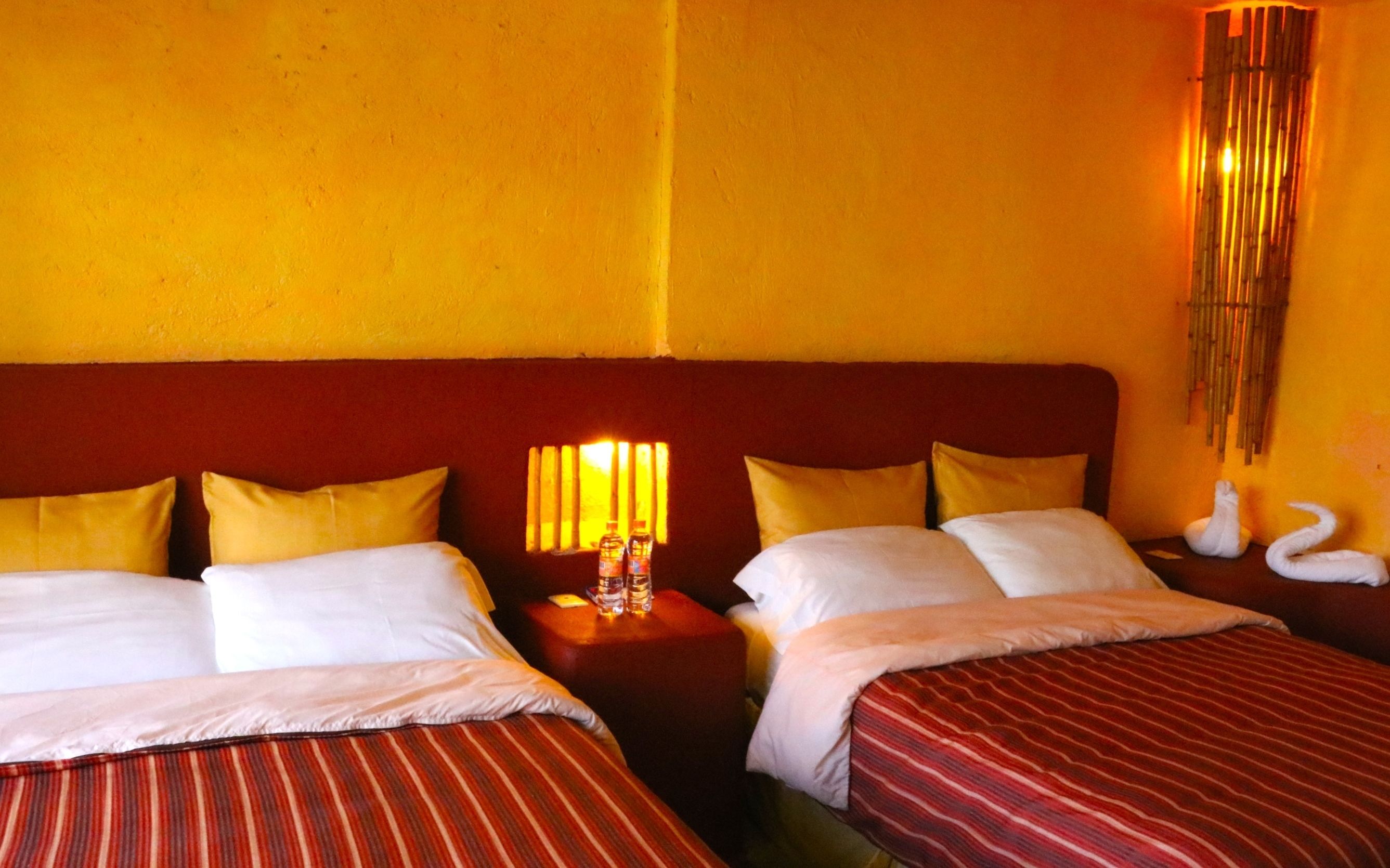 A hotel room with two beds, each with white linens and striped red bedding. There is a nightstand with a lamp and two bottles of water. The walls are painted yellow, and there is a decorative wall hanging with swans.