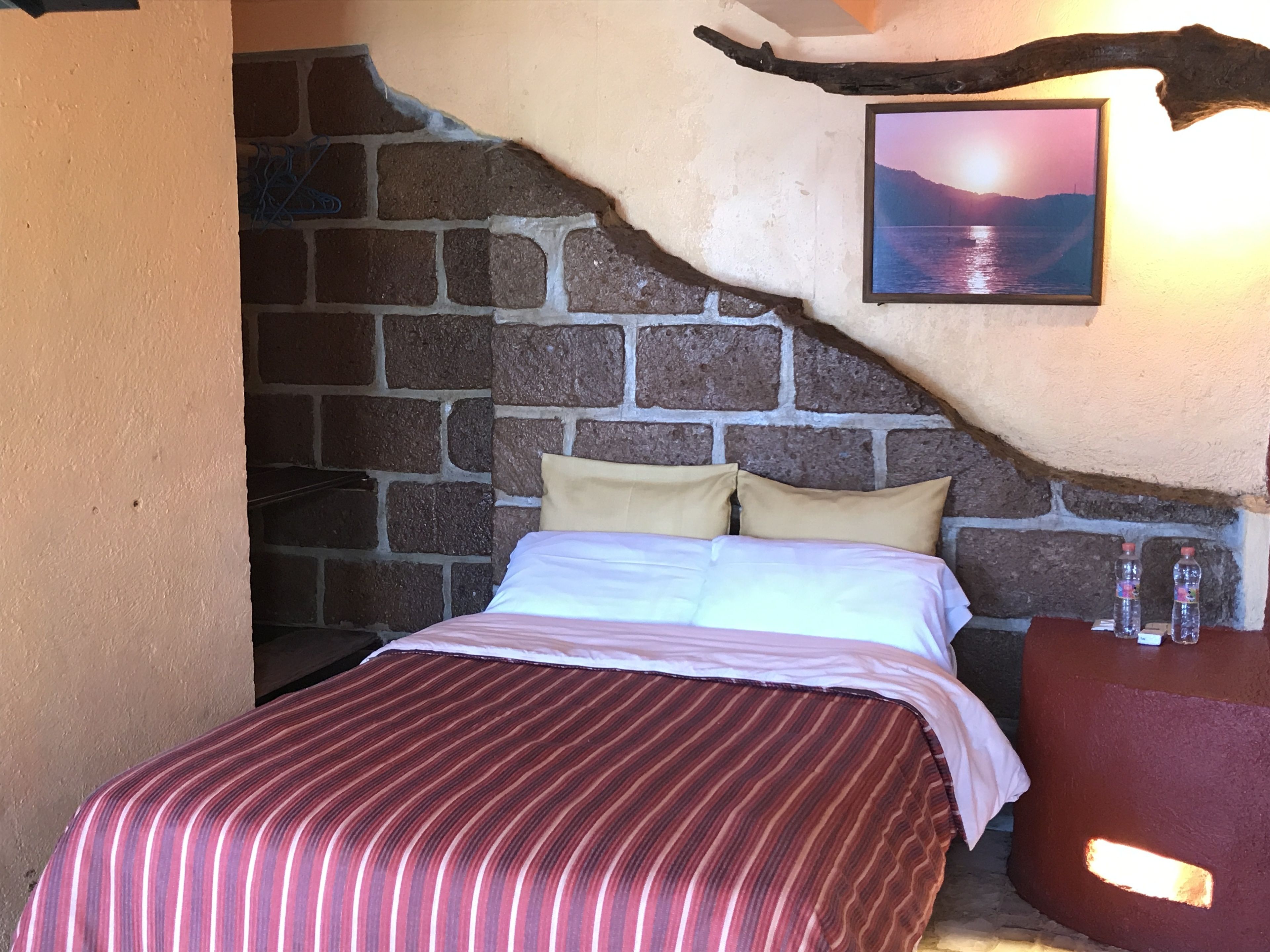 A bed with a striped bedspread, a headboard, and a pillow. There is a picture on the wall and a small table with bottles.