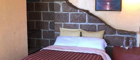 A bed with a striped bedspread, a headboard, and a pillow. There is a picture on the wall and a small table with bottles.