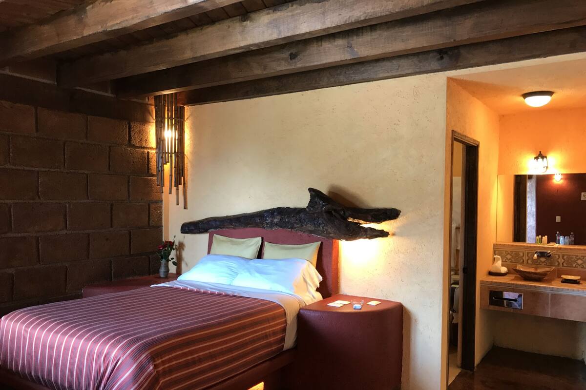 A room with a bed, a nightstand, a wooden headboard, a wall-mounted light fixture, and a bathroom with a mirror and sink.