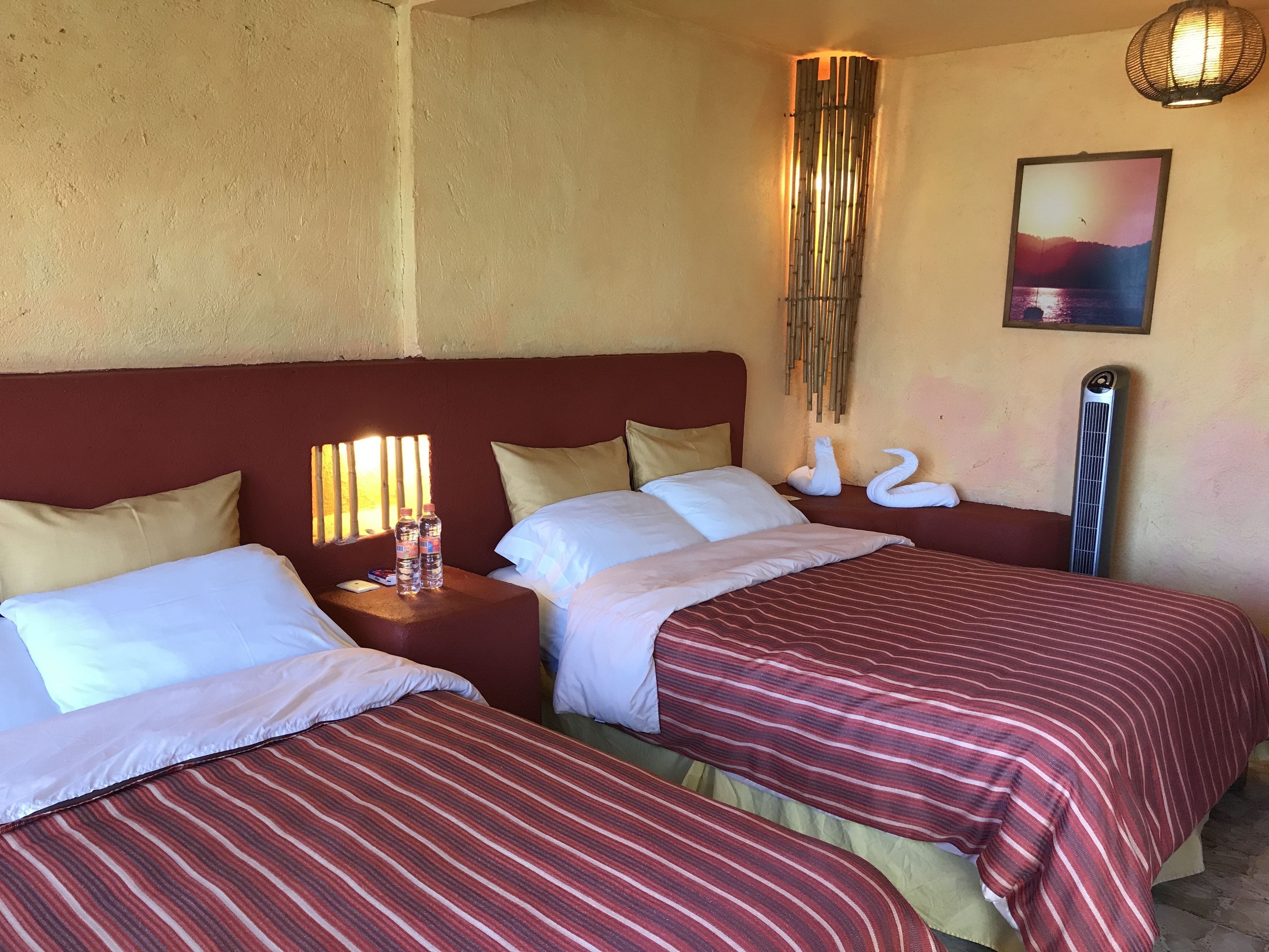 A hotel room with two beds, each with striped bedding, a nightstand with water bottles, a wall-mounted heater, and a framed picture on the wall.