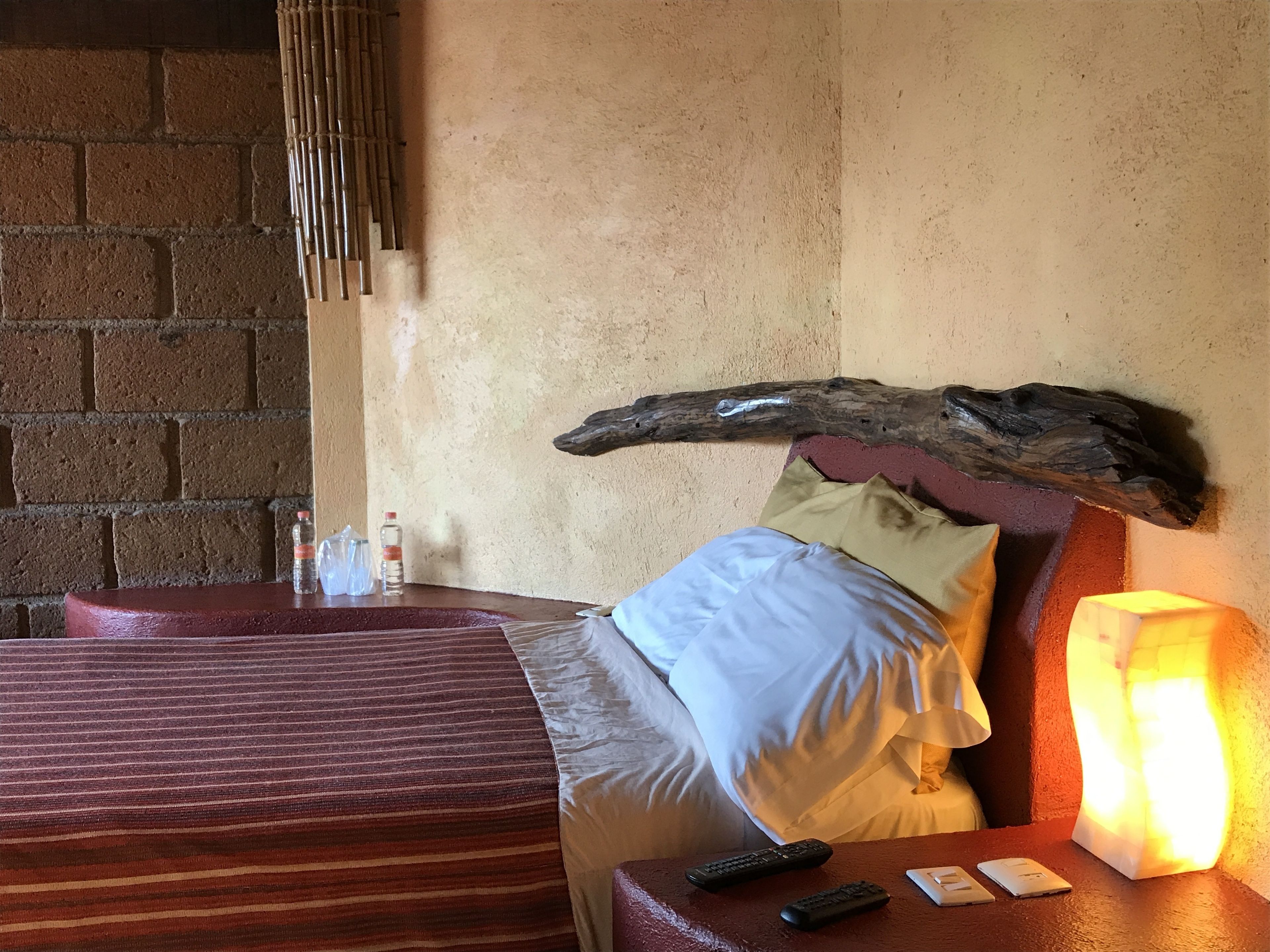 Una habitación de hotel con cama, cabecera de madera, mesita de noche con lámpara y pared de ladrillo.