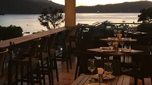 Un área de comedor al aire libre con mesas y sillas, con vistas a un cuerpo de agua y colinas.