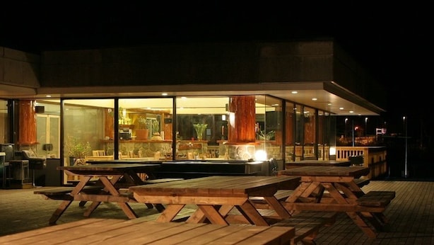 Een terras met houten tafels en stoelen, grote ramen en een stenen muur.