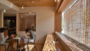 A modern dining area with wooden furniture, a long table, and a view through bamboo blinds.