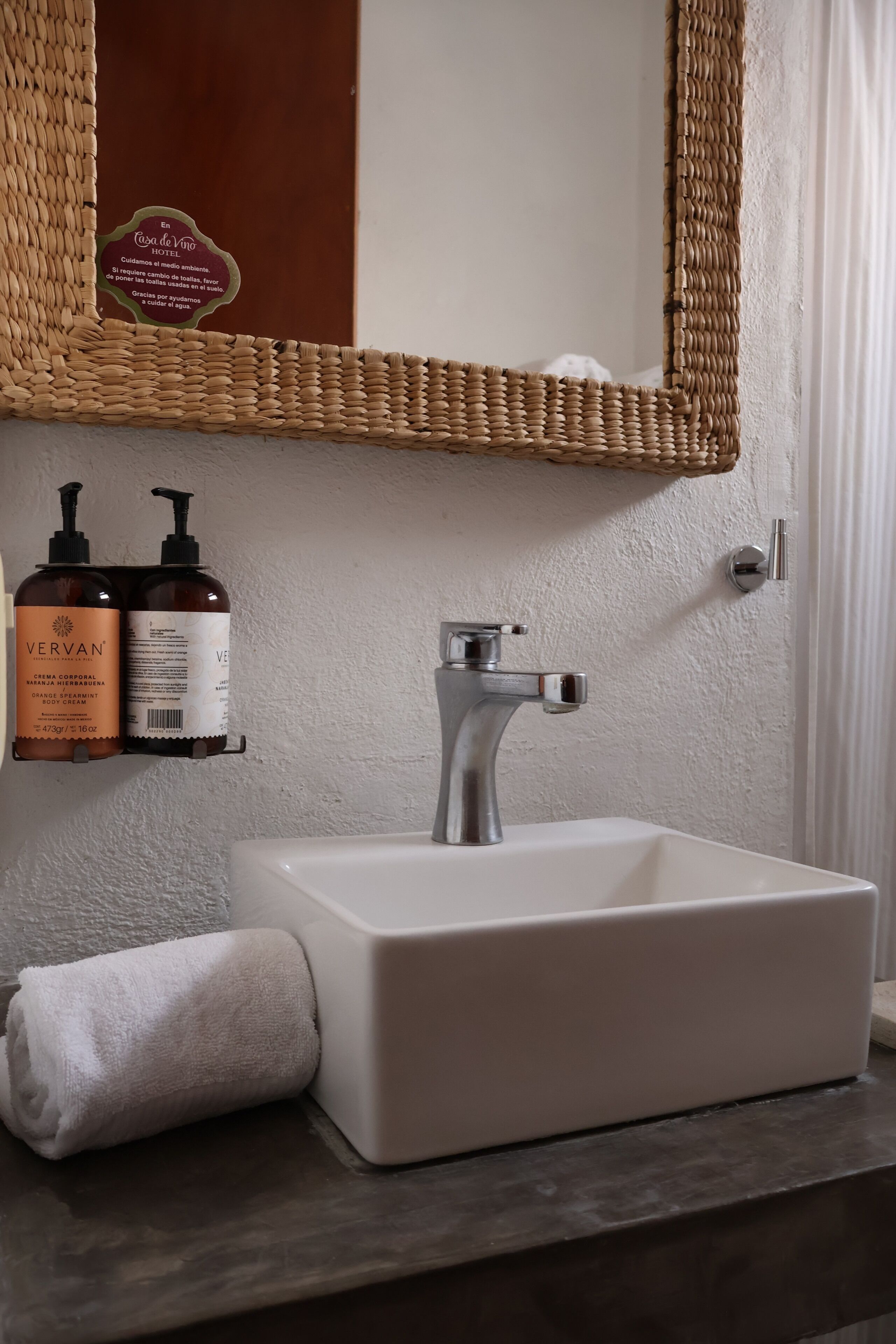 A bathroom with a white sink, a chrome faucet, a woven basket shelf, and two bottles of Vervan body cream.