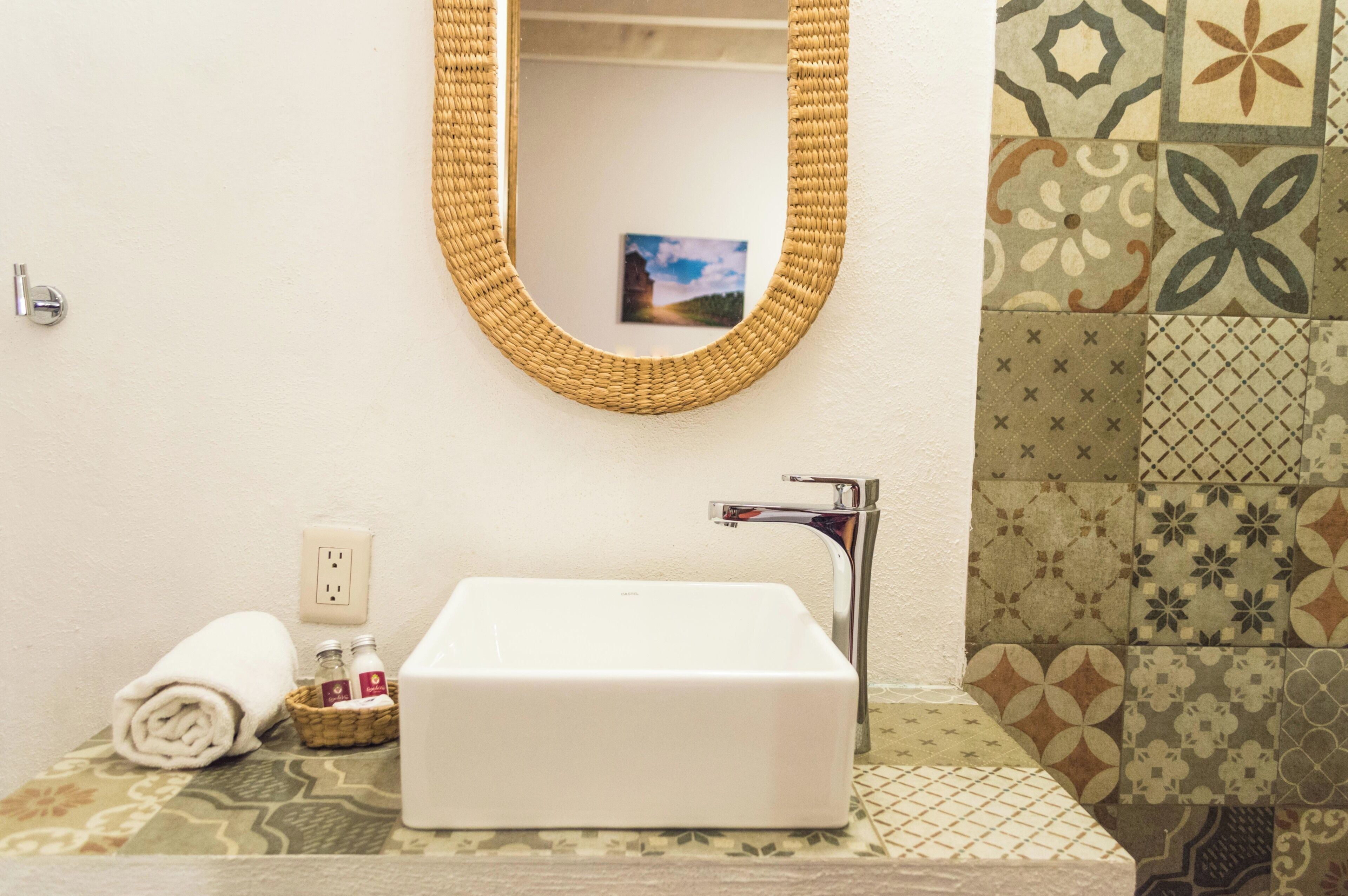 A bathroom with a white sink, a woven mirror, a towel, and a bottle.