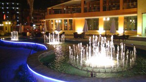 Un espace extérieur agrémenté d’une fontaine illuminée la nuit, avec de nombreux jets d’eau, entouré d’une piscine et d’un bâtiment doté d’une terrasse.