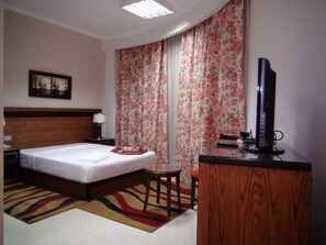 A hotel room with a bed, a desk, a chair, a television, a nightstand, a lamp, a painting on the wall, and a window with floral curtains.