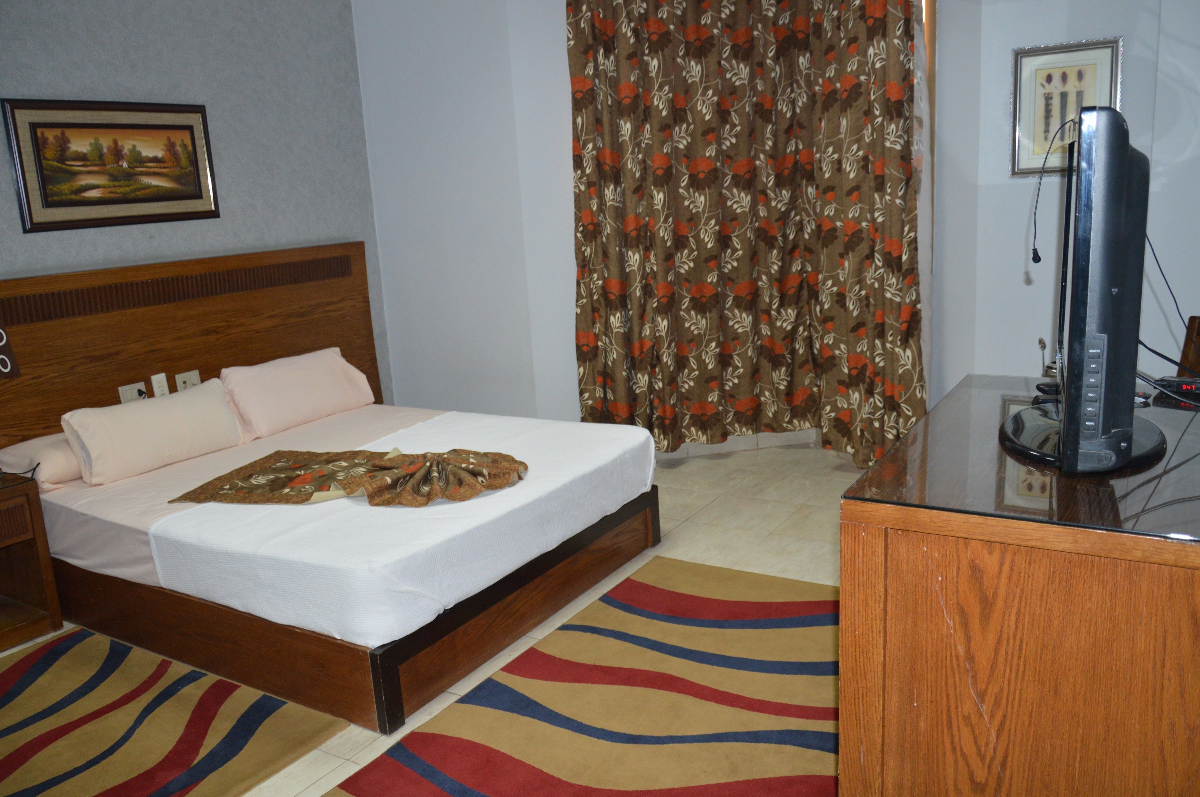 A hotel room with a wooden bed, a flat-screen TV, a desk, and a patterned curtain.