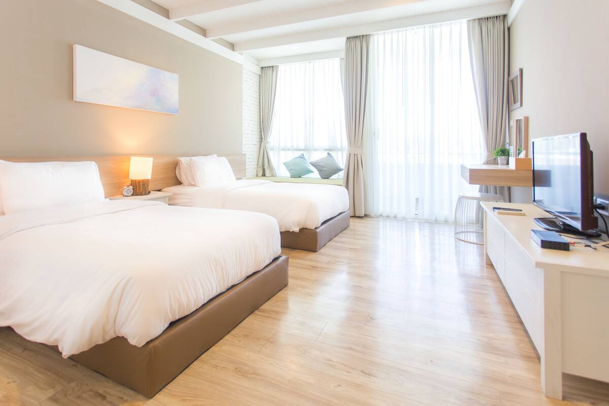 A modern hotel room with two beds, a desk, a TV, and a large window with curtains.