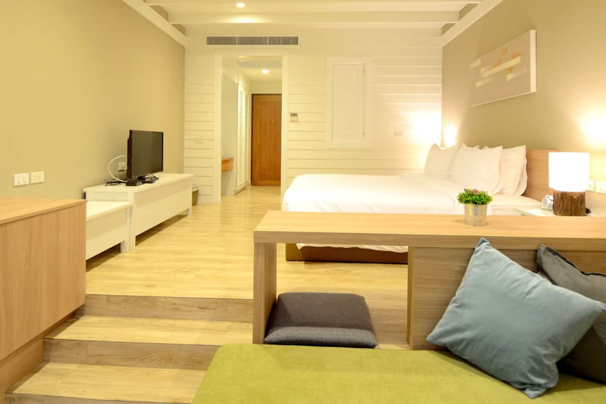 A modern hotel room with a wooden floor, a bed with white linens, a desk with a computer, and a small green plant.