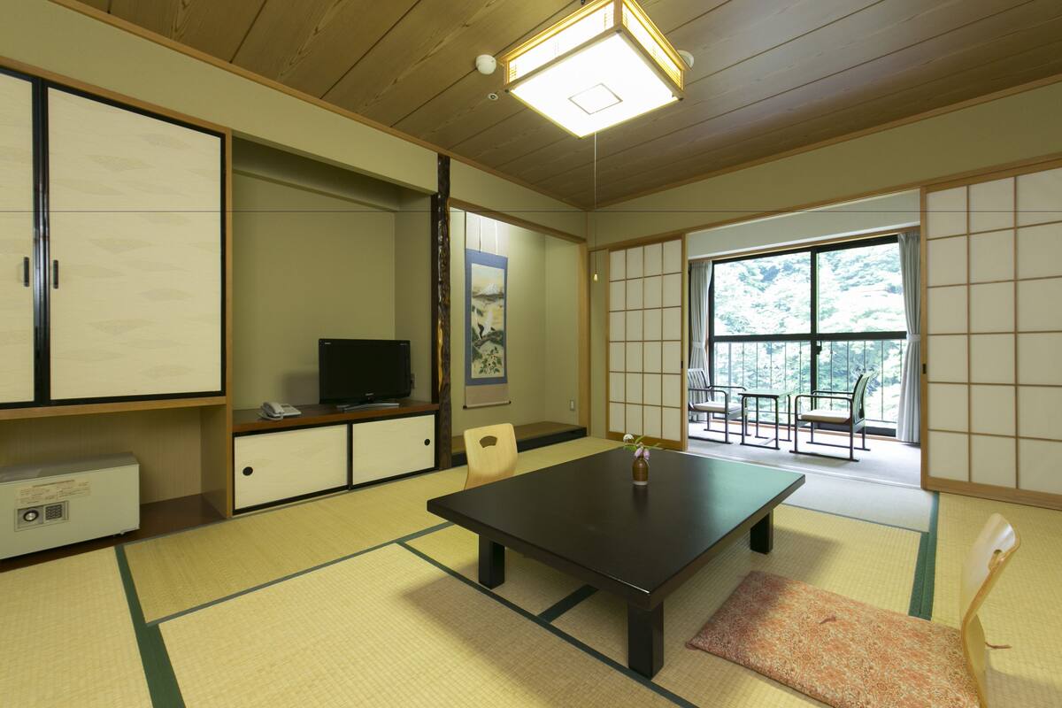 A traditional Japanese room with tatami flooring, a low table, chairs, a television, and a large window with a view of trees.