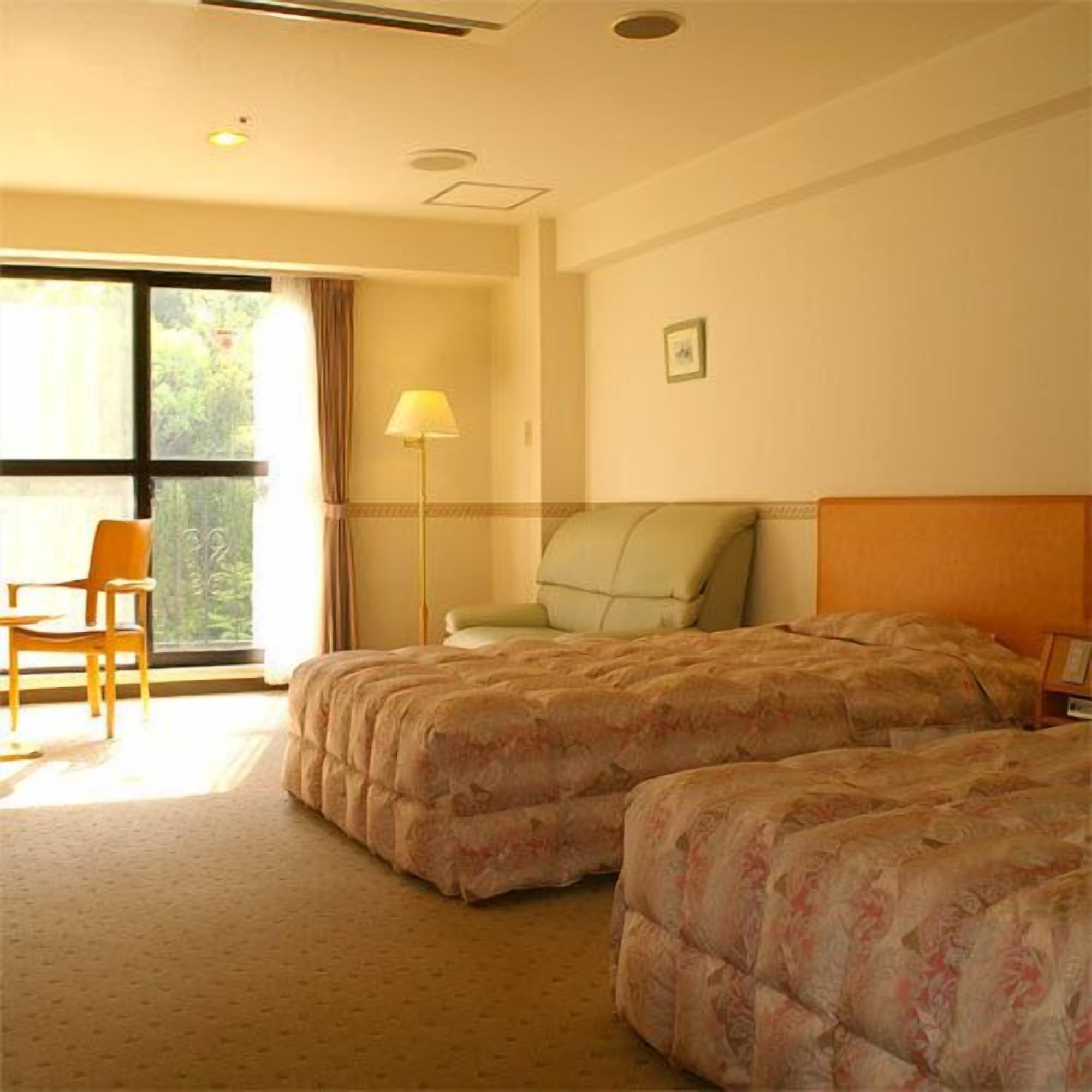 Quarto de hotel com duas camas, uma cadeira, um abajur e uma janela com cortinas.
