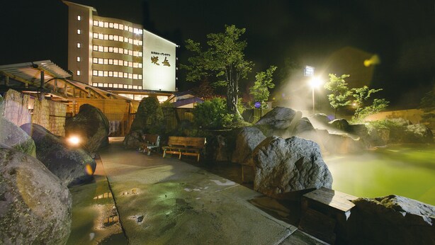 A traditional Japanese onsen with a wooden deck, large rocks, and a building in the background. - Okuhida Garden Hotel Yakedake (Takayama)
