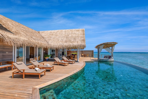 A resort with a wooden deck, lounge chairs, a pool, and a thatched-roof structure.