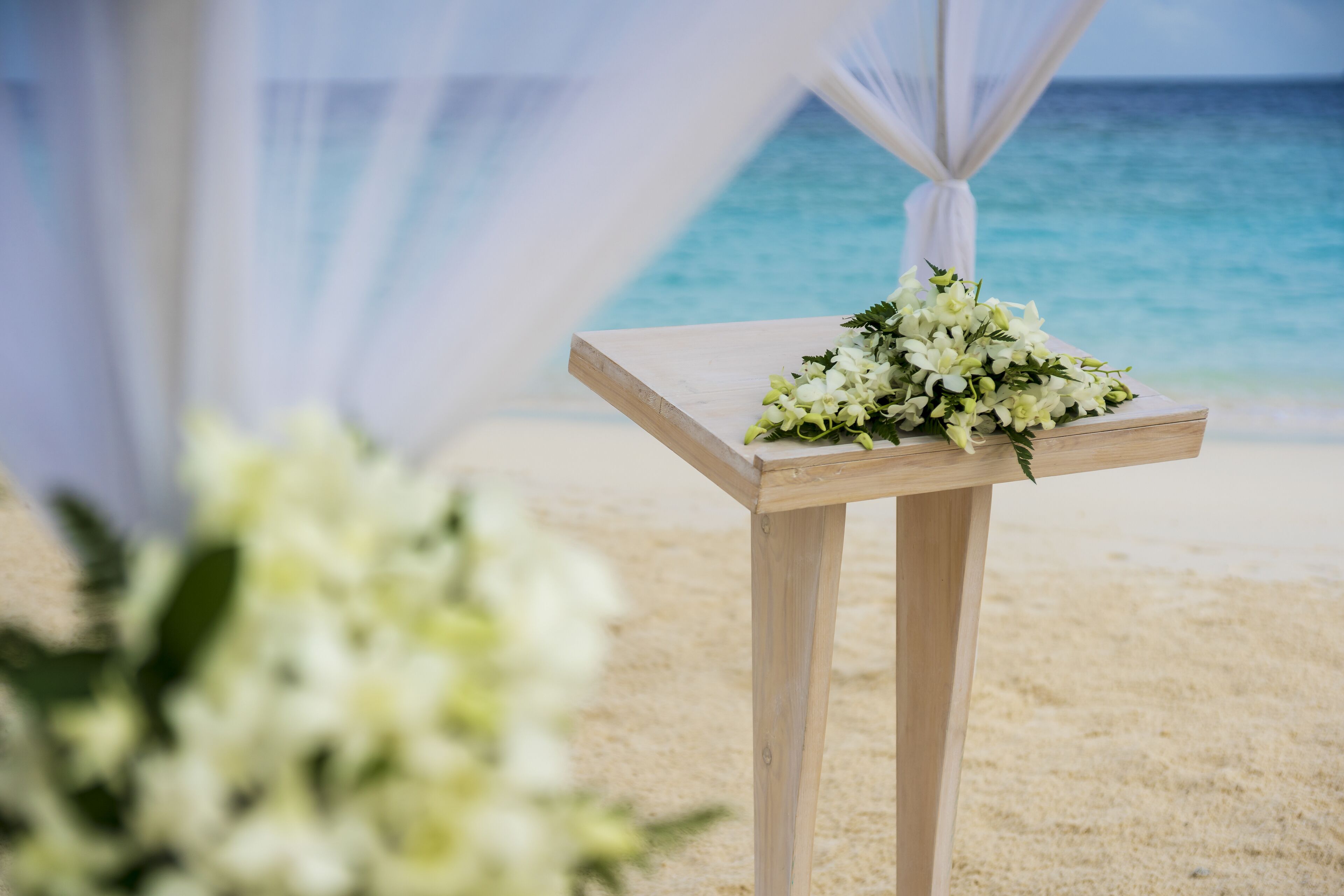 A wooden table with a floral arrangement on a beach with a clear view of the ocean.