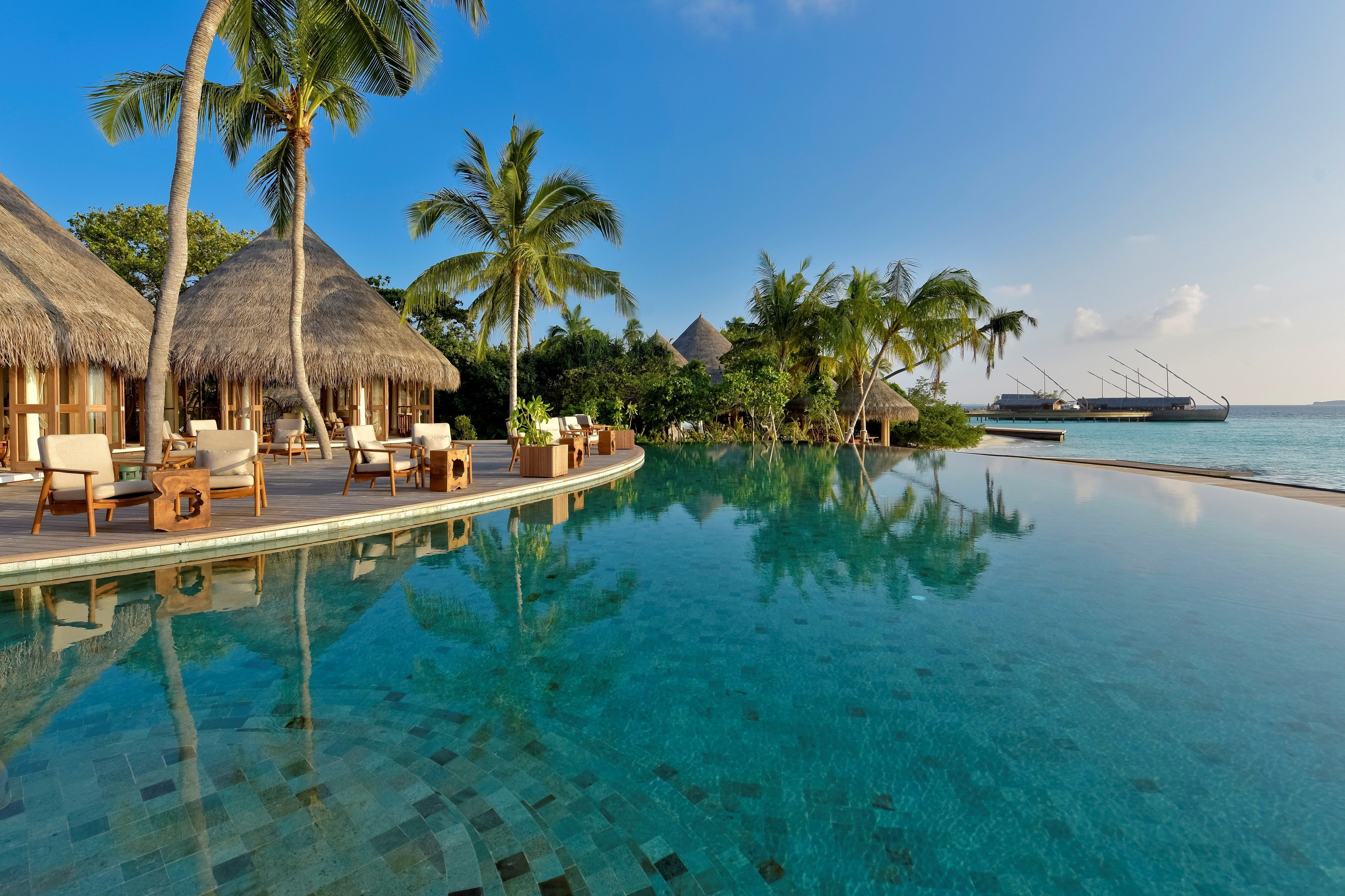 An infinity pool with clear blue water, surrounded by lounge chairs and palm trees, with a boat docked in the background.