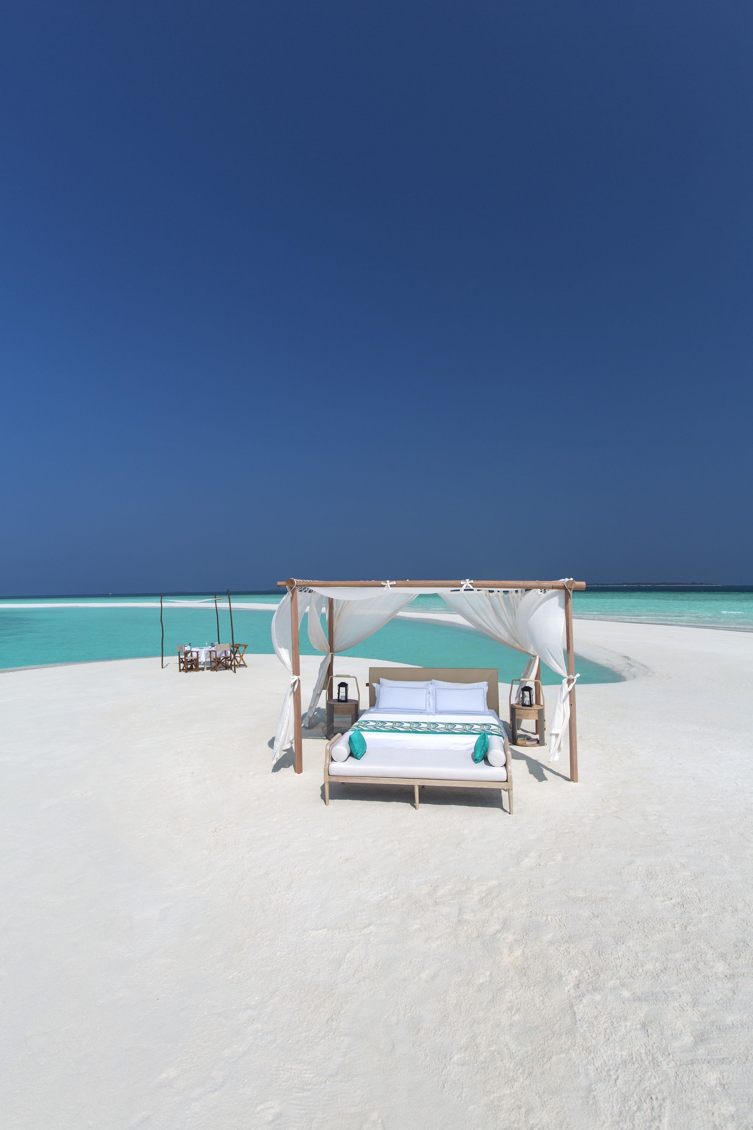 A beach cabana with a bed, a table, and chairs, overlooking a clear turquoise sea.