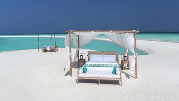 A beach cabana with a bed, a table, and chairs, overlooking a clear turquoise sea.