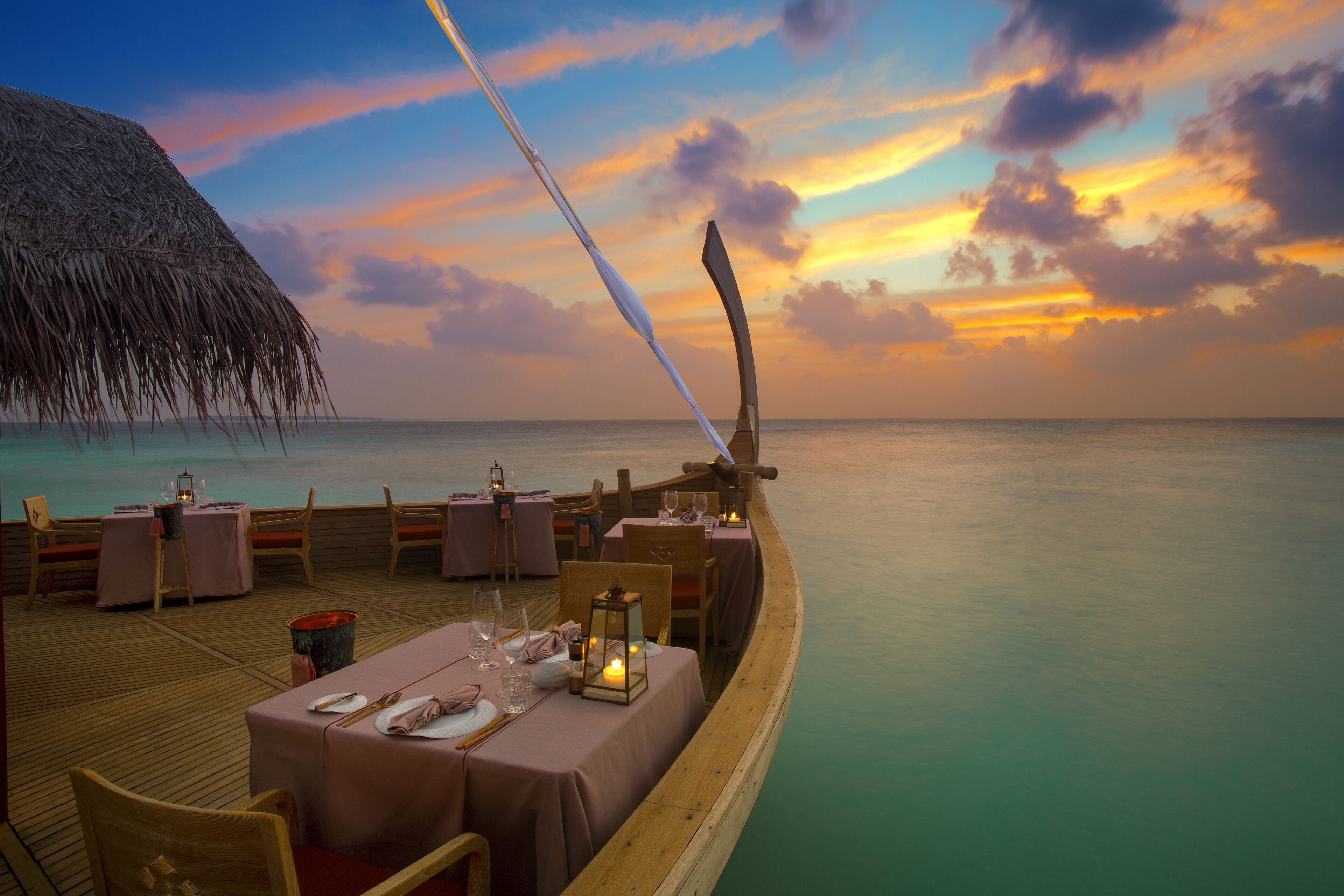 A dining area on a boat with tables set for two, overlooking a calm sea and a sunset.
