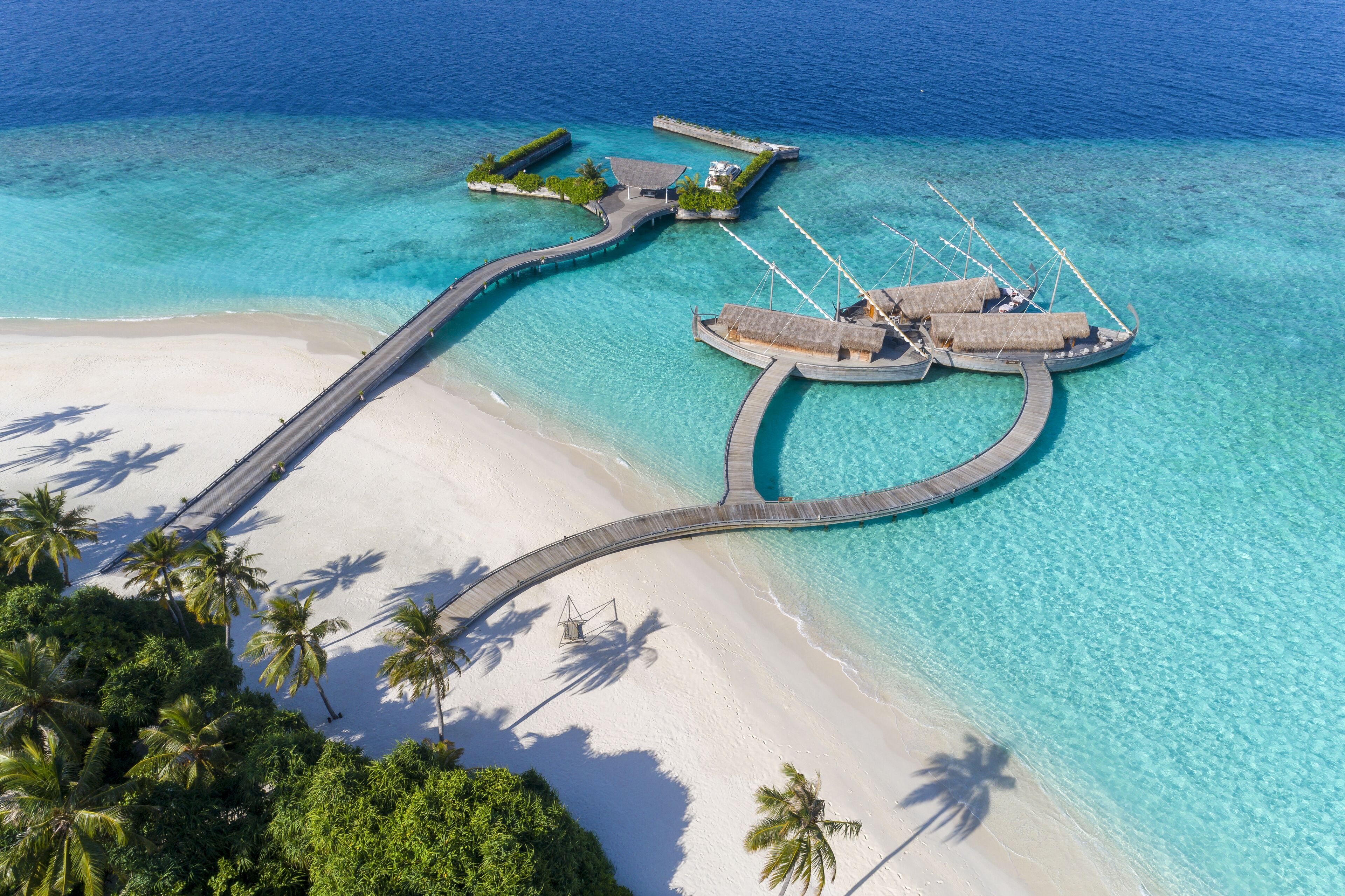 An aerial view of a resort with overwater bungalows connected by a bridge to a sandy beach and palm trees.
