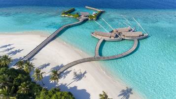 An aerial view of a resort with overwater bungalows connected by a bridge to a sandy beach and palm trees.