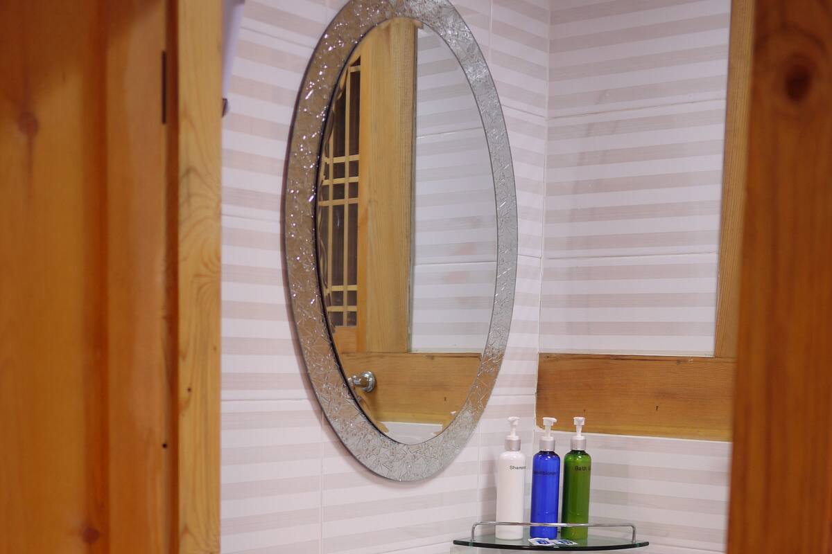 Un baño con puerta de madera, un lavabo blanco con grifo plateado, repisas de vidrio con artículos de aseo, y un espejo con marco decorativo.