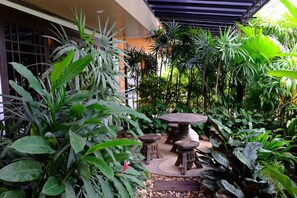Um jardim com uma mesa redonda e banquinhos, rodeado por diversas plantas tropicais.