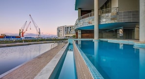 A swimming pool adjacent to a building with a balcony, a view of the water, and construction cranes in the background. - 702 Canal Quays (Cape Town)