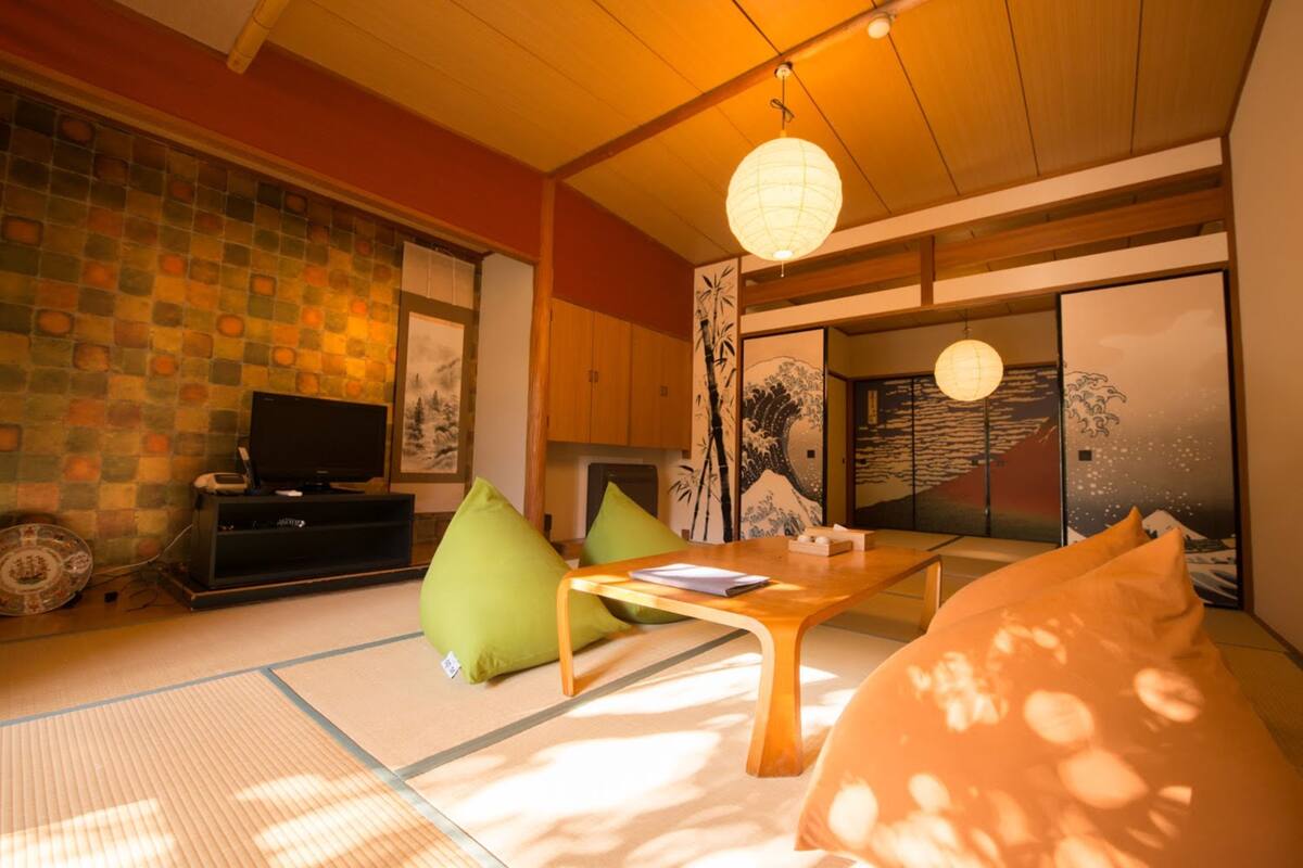 A traditional Japanese room with tatami flooring, a low wooden table, green cushions, a television, and sliding doors with artwork.