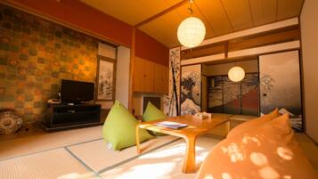 A traditional Japanese room with tatami flooring, a low wooden table, green cushions, a television, and sliding doors with artwork.