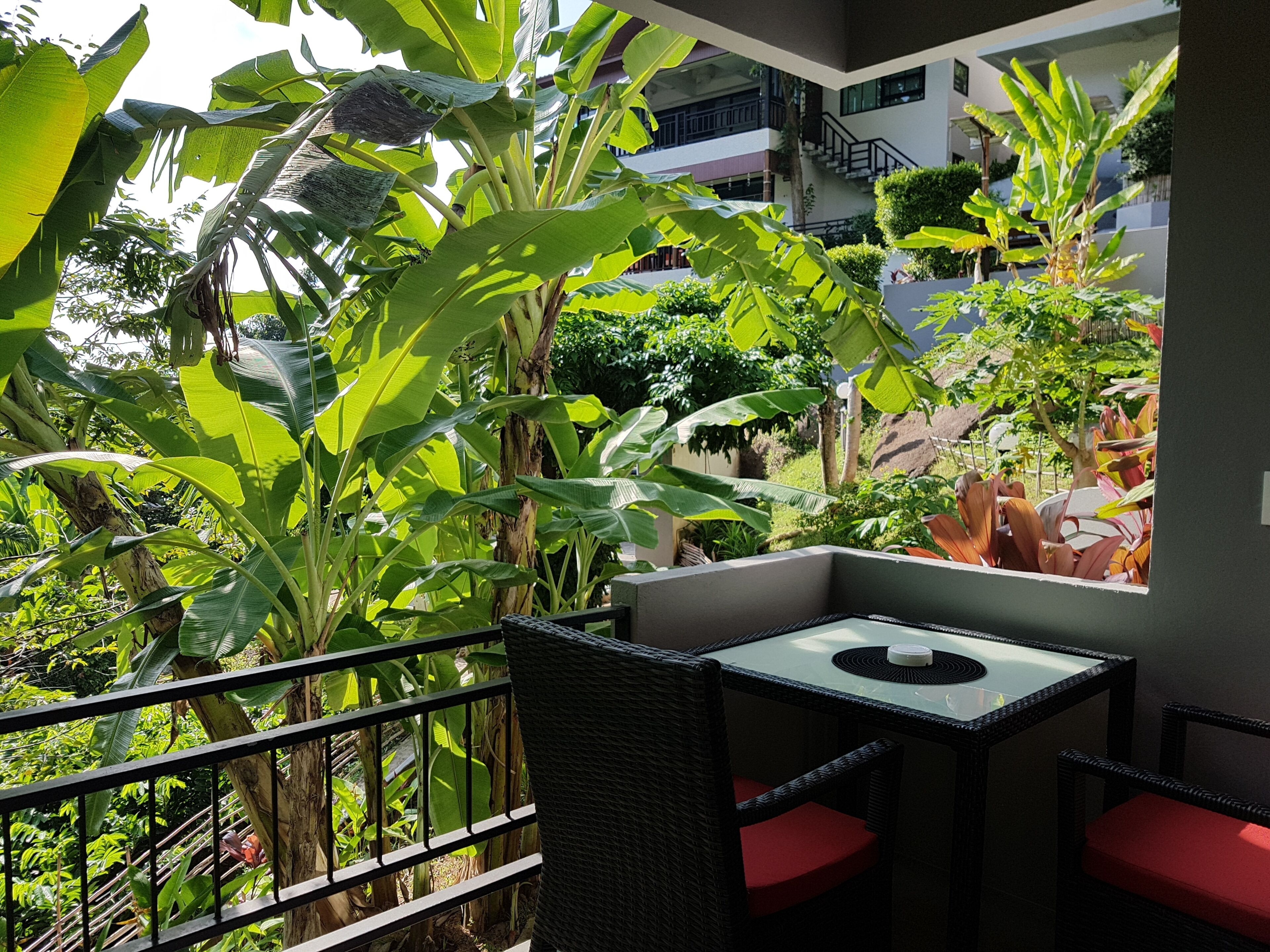 Un balcón con mesa y sillas, rodeado de exuberante vegetación tropical y vistas a un edificio moderno.