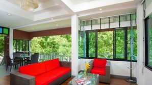 A modern living room with a red sofa, a glass coffee table, and a dining area with a table and chairs. Large windows offer a view of greenery outside.