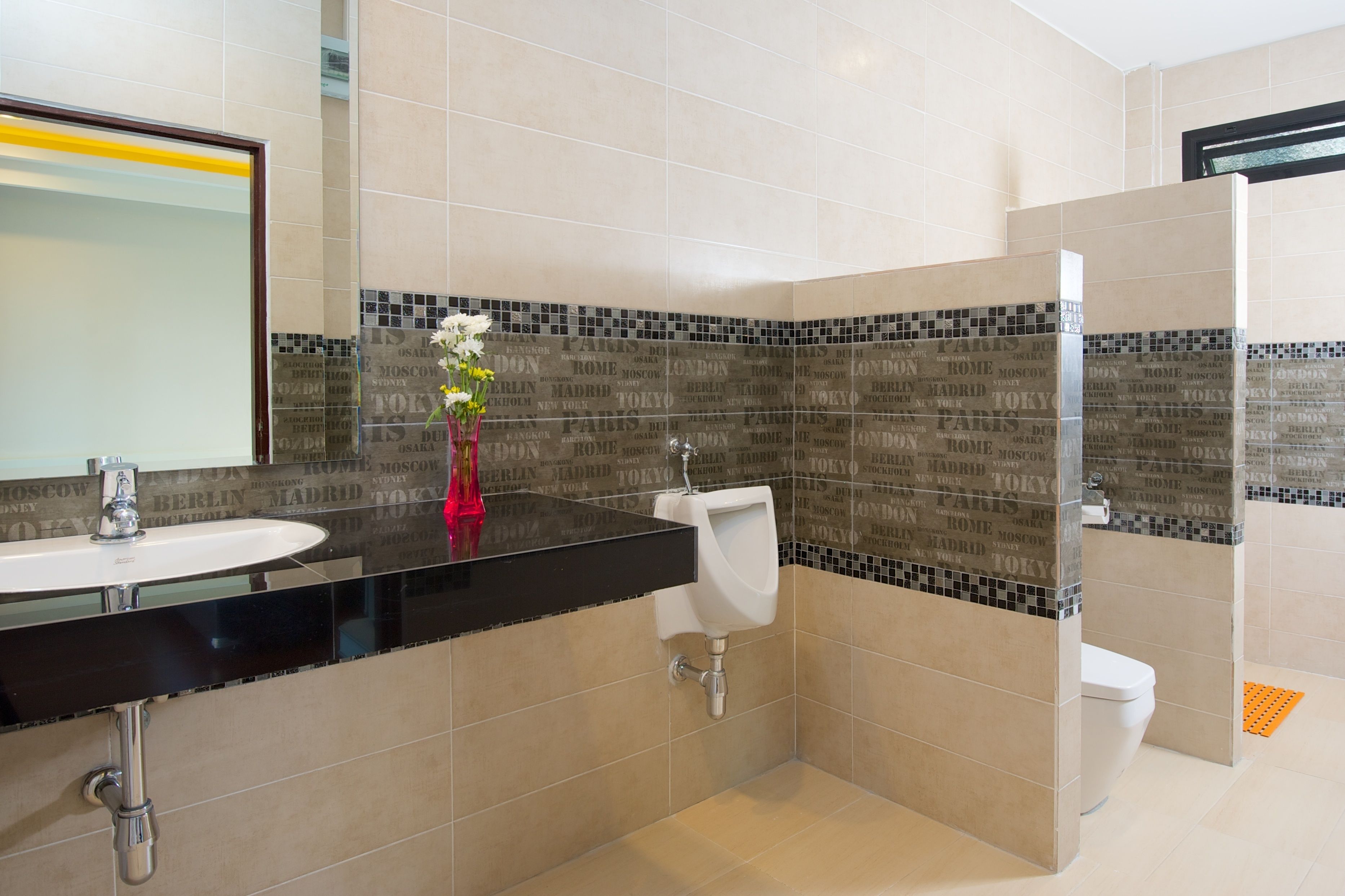 Un baño moderno con lavamanos, espejo e inodoro. Las paredes tienen un diseño de baldosas con nombres de ciudades.
