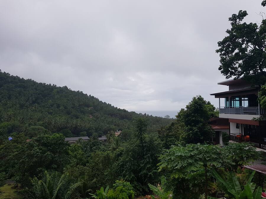 A multi-story building with a balcony, surrounded by dense tropical vegetation and a hilly landscape.