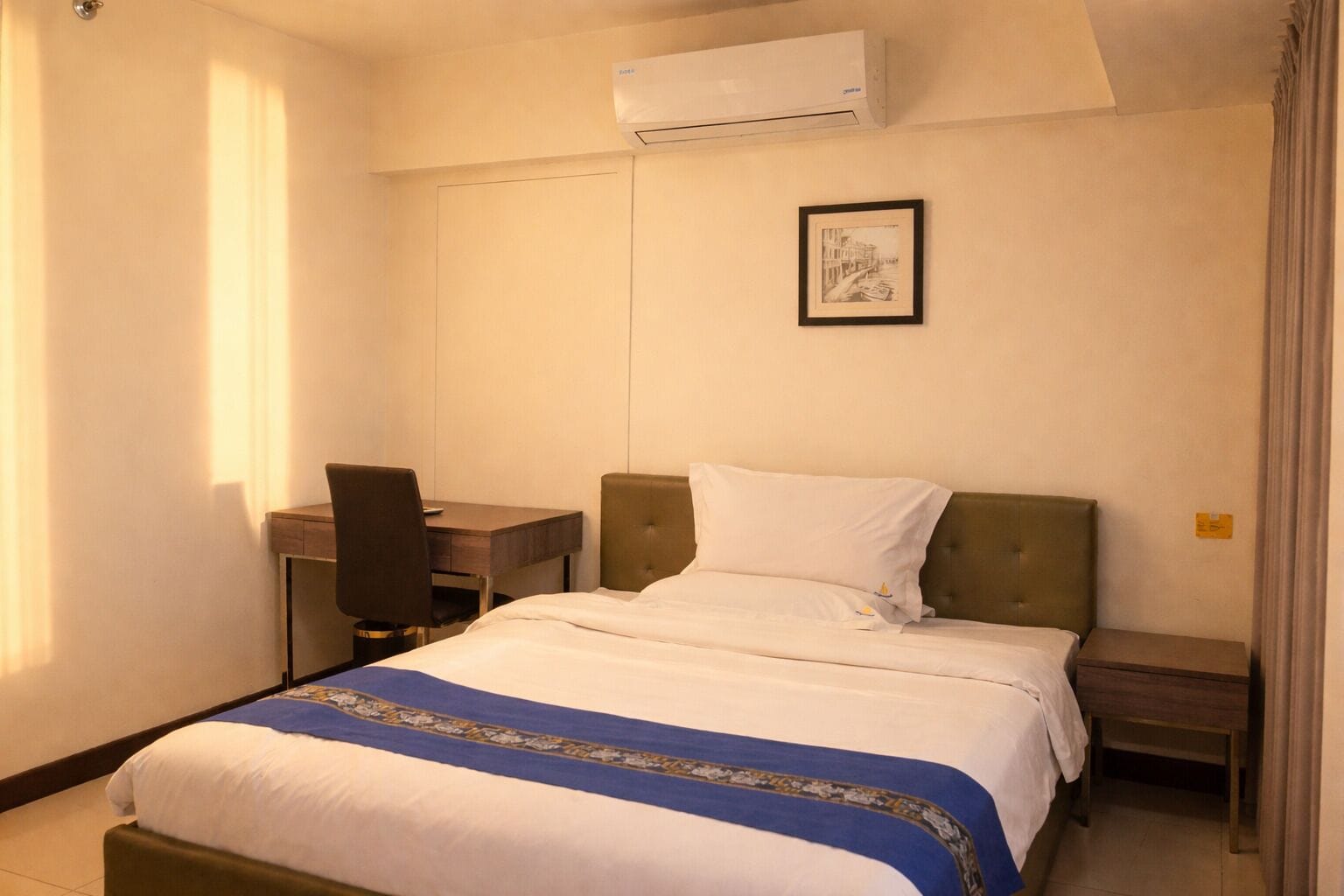 Una habitación de hotel con cama, escritorio, silla, aire acondicionado y un cuadro en la pared.
