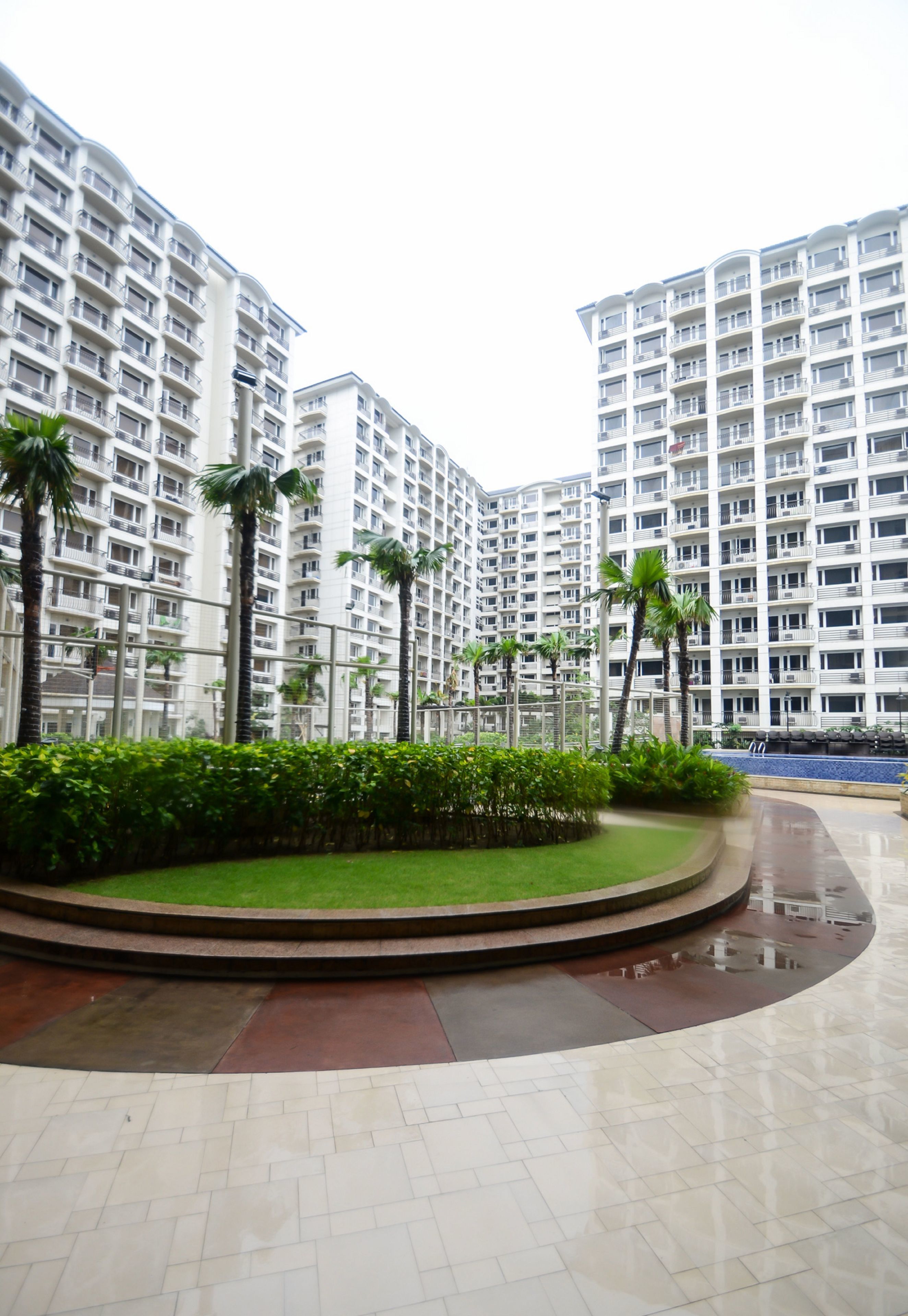 A modern residential building with multiple balconies, a well-maintained garden area, and a reflective tiled floor.