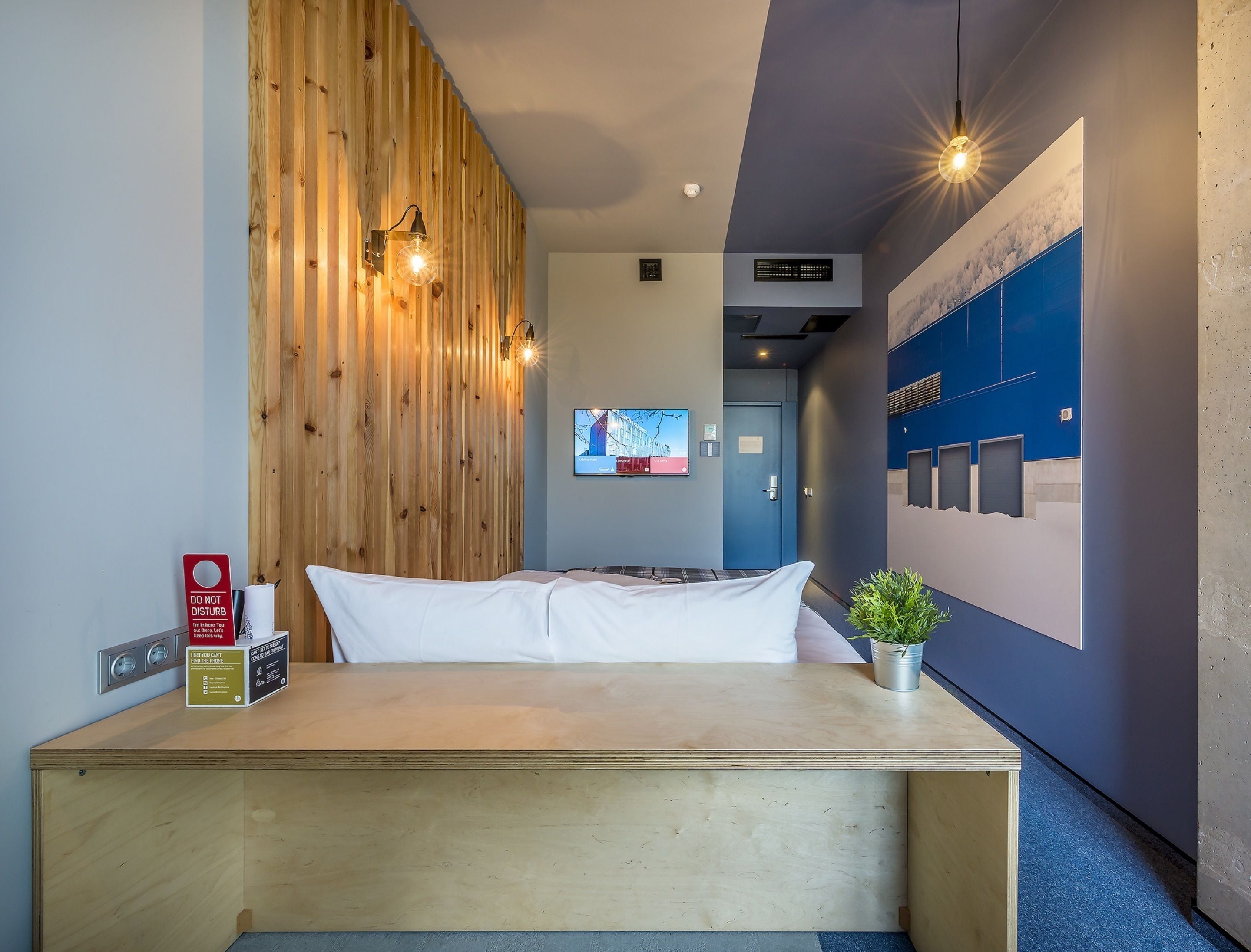 Hotelkamer met een houten accentmuur, een bed met wit beddengoed, een wandgemonteerde televisie en een kleine potplant.