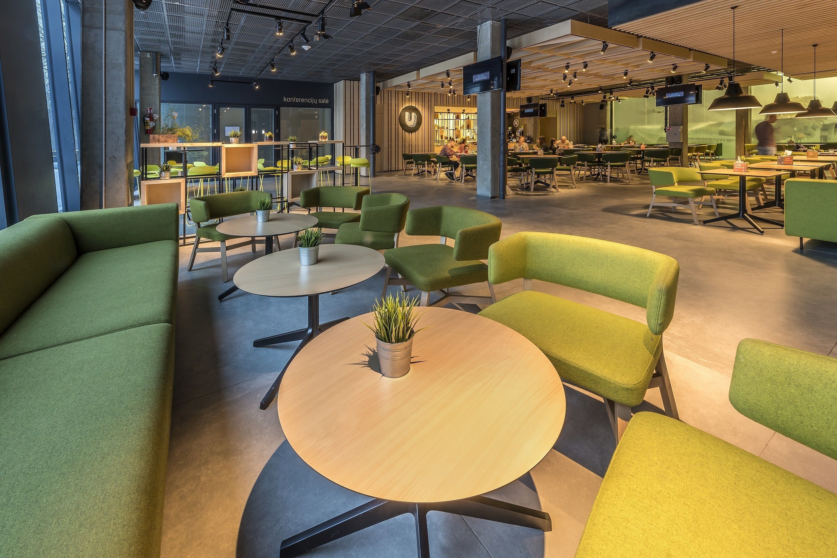 A modern cafe with green seating, round tables, and a large TV screen.