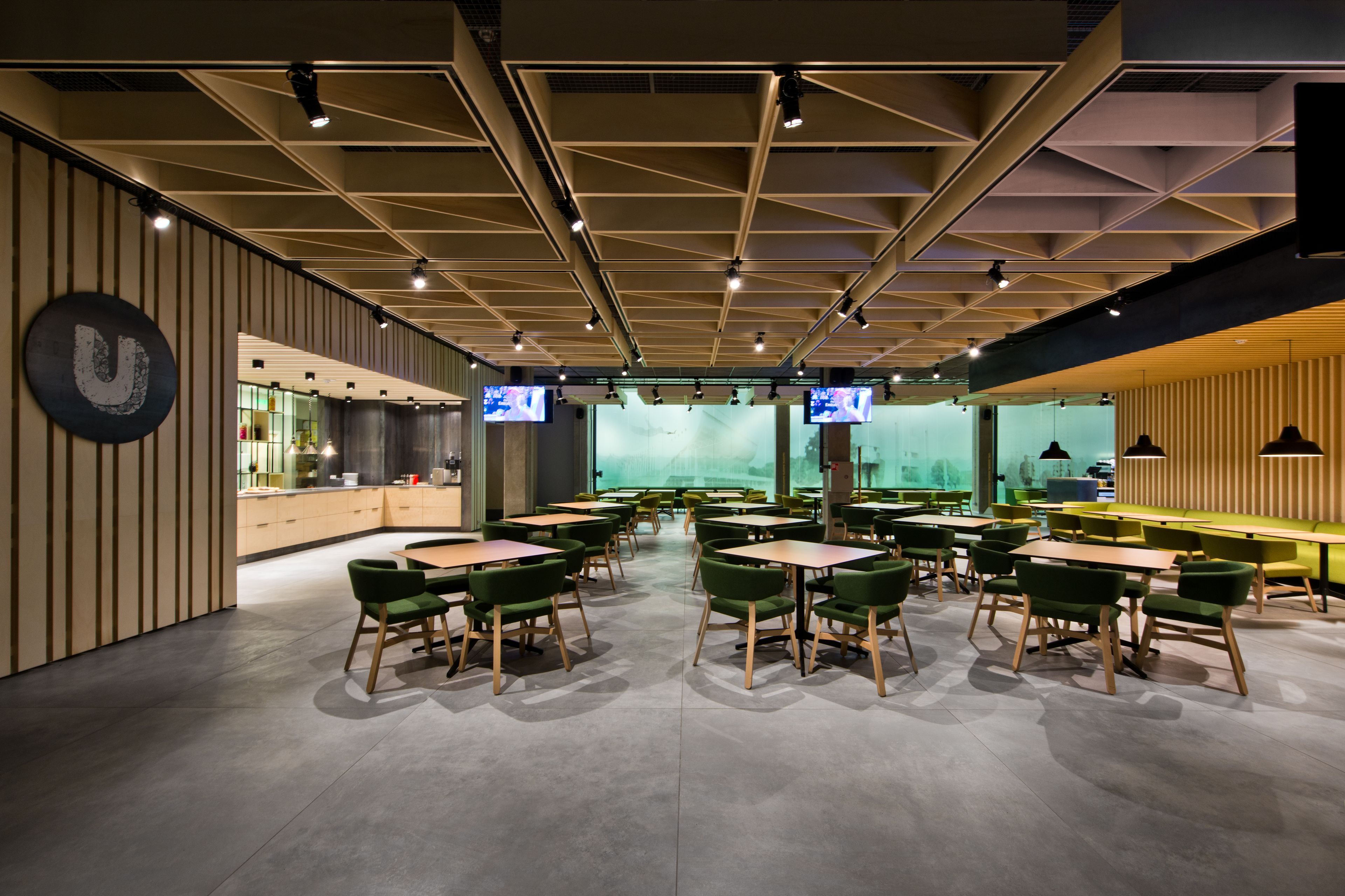 Un restaurant moderne doté d’un plafond au design géométrique, de chaises recouvertes de tissu vert et de plusieurs téléviseurs fixés aux murs.
