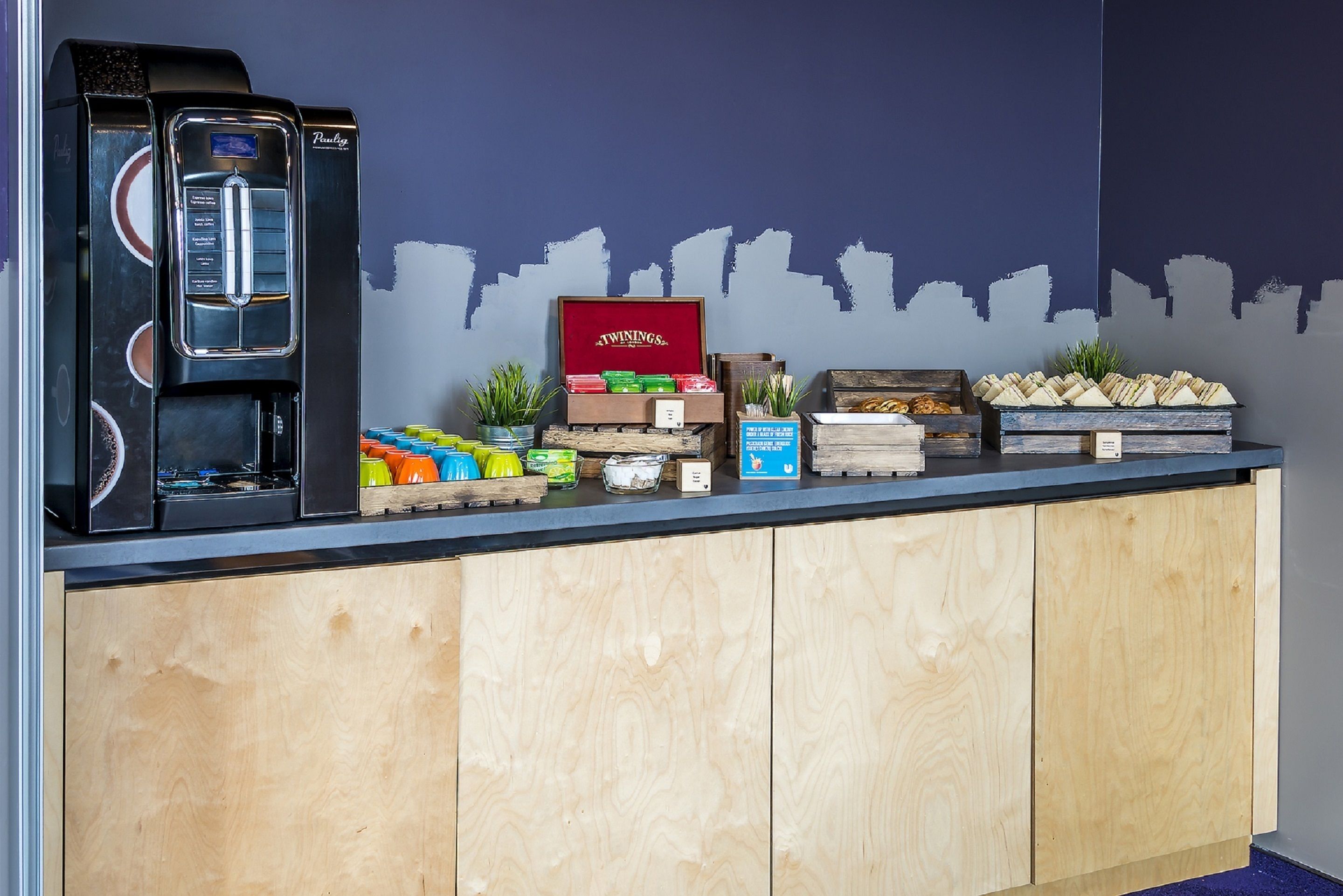 Una barra con una máquina de café, cajas de té Twinings, aperitivos y un mural de un paisaje urbano.