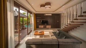 A modern living room with a sofa, coffee table, and TV. There is a staircase with wooden steps and a glass door leading outside. - Sotis Villa Canggu (Canggu)